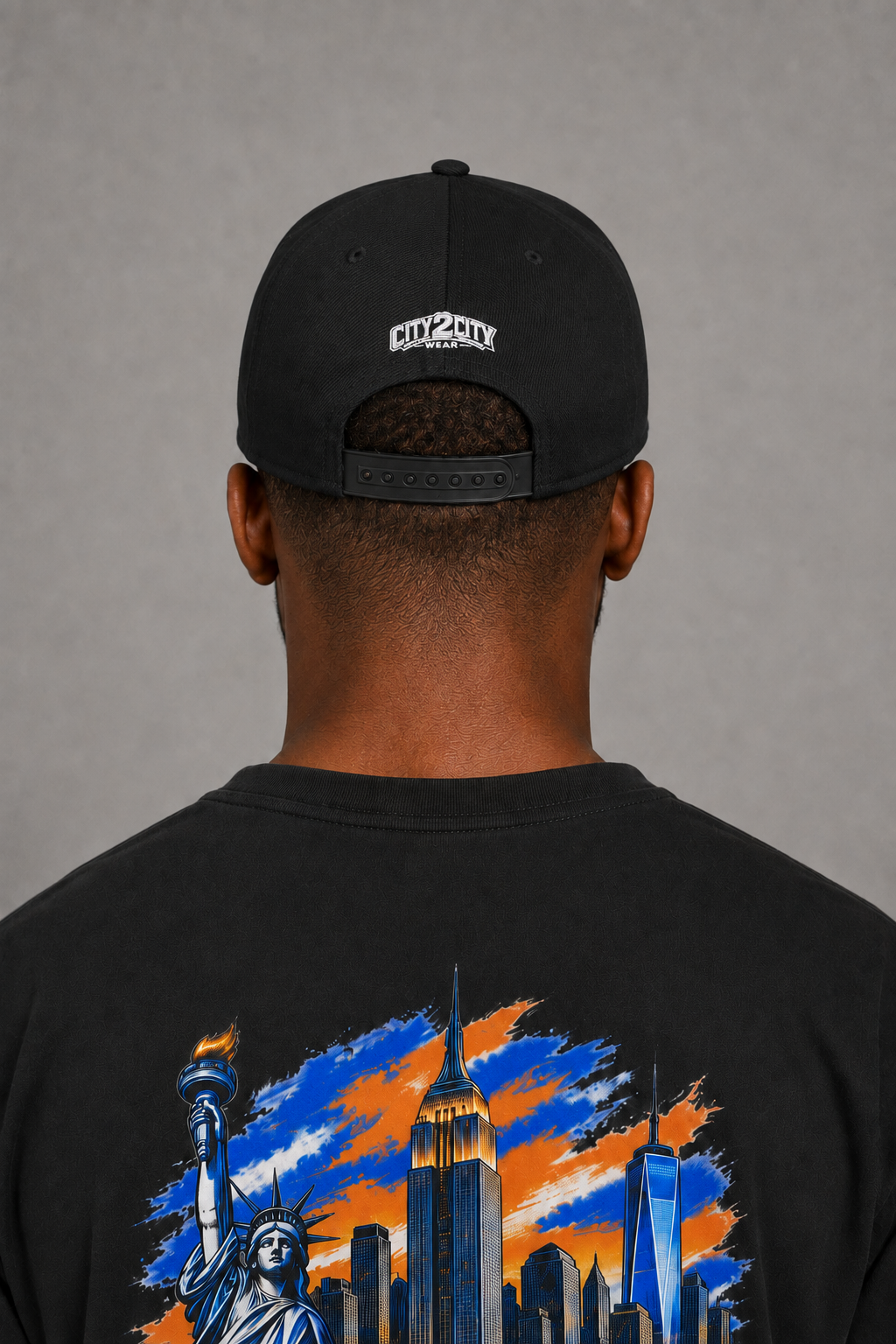 New York City Concrete Jungle Black Embroidered Snapback Hat — close-up rear model view showing white City 2 City Wear embroidery, black adjustable snap closure, and blue-orange NYC skyline artwork on the black T-shirt back | City 2 City Wear