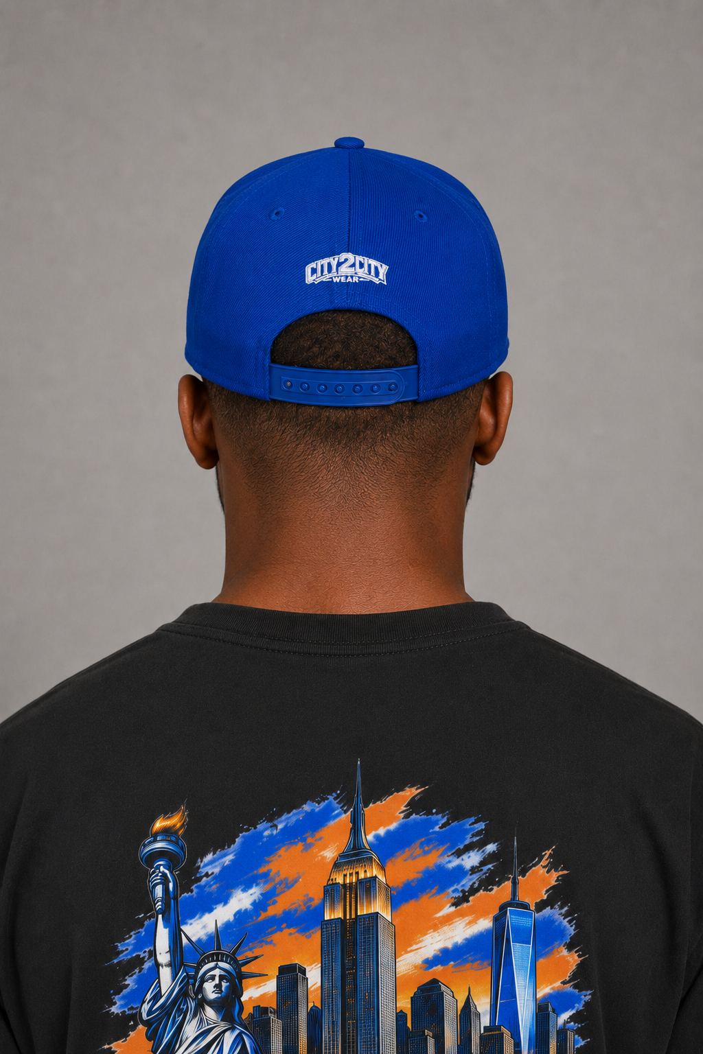 NYC Concrete Jungle Royal Blue Embroidered Snapback Hat — close-up rear New York City model view showing white City 2 City Wear embroidery, royal-blue adjustable snap closure, and blue-orange NYC skyline artwork on the black T-shirt back | City 2 City Wear